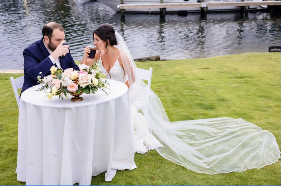 venues pensacola fl with bride and groom sharing a private moment together sipping cocktails on the lawn she in white and he in blue suit and she has a very long veil sitting at white linen table and pink and ivory florals