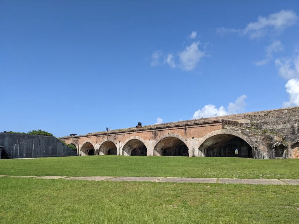 This stunning photo features a historic fort with a series of elegant brick arches, set against a vibrant green lawn and a clear blue sky with scattered clouds. The architectural beauty of the structure, combined with its open, scenic surroundings, makes it a captivating backdrop for special events. This couple touring this location is want to go back there after their ceremony at the location which is one of the top wedding places in Pensacola, FL, offering a unique blend of history and natural beauty for a memorable wedding ceremony.
