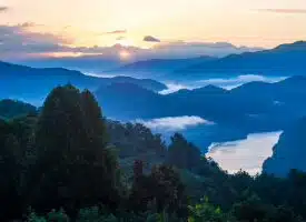 One of the Road Trips - This breathtaking photo captures a serene mountain landscape at sunset, with the sun casting a warm golden glow over rolling blue ridges shrouded in mist. A tranquil river winds through the lush, forested valley, framed by towering trees in the foreground. The peaceful and picturesque setting makes it an ideal choice among wedding places Pensacola, offering a stunning natural backdrop for a memorable outdoor wedding ceremony.