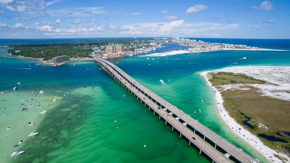 This vibrant aerial photo showcases a stunning coastal scene with a long bridge stretching over crystal-clear turquoise waters, connecting a lively beach area to a bustling island town. The image features sandy shores dotted with boats and swimmers, vibrant green waters, and a backdrop of modern buildings under a bright blue sky with scattered clouds. The newly engaged couple are considering their wedding receptions in Pensacola, FL, and this picturesque waterfront setting offers an ideal venue for a memorable time after they get married in Pensacola.
