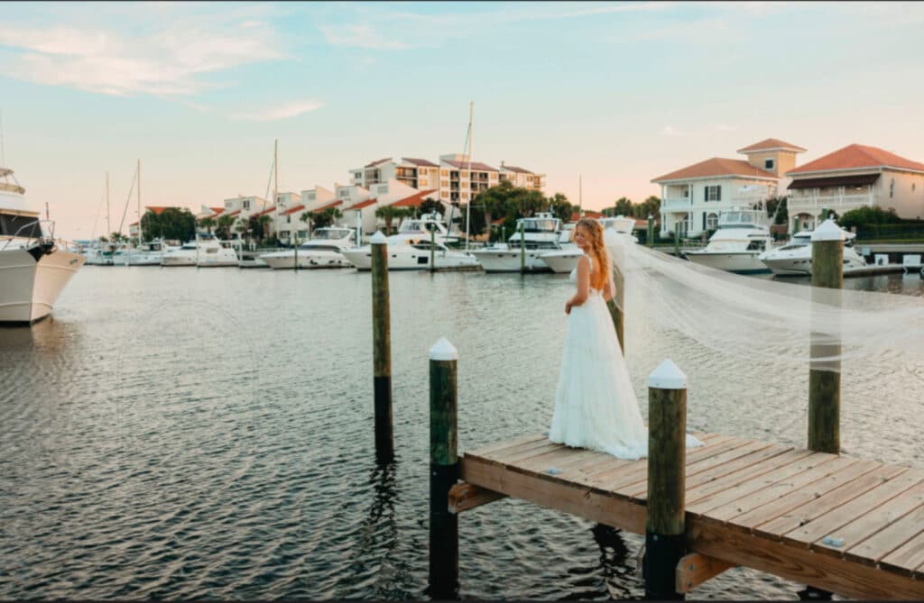 Pensacola wedding venues and this one is Palafox Wharf Waterfront Wedding Venue with the bride on the dock wearing her white bride’s dress watching the beginning of a gorgeous sunset across the waterfront with yachts, boats, and upscale luxury homes and she used a wedding day checklist with her planner