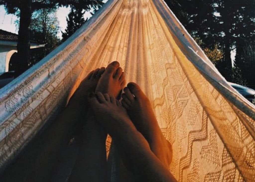 This cozy photo captures a romantic moment with two pairs of feet resting side by side in a beautifully patterned hammock, bathed in the warm golden light of sunset. The scene is framed by lush trees and a glimpse of a house, creating a serene and intimate atmosphere. The newly engaged couple are considering their wedding venues in Pensacola, FL, and this tranquil outdoor setting offers a charming and relaxed option to dream about their special day.
