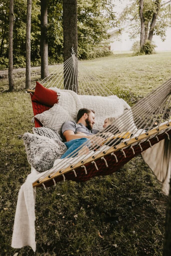 This heartwarming photo depicts a couple relaxing together in a cozy hammock nestled among tall trees on a lush green lawn, surrounded by a serene natural setting. The hammock is adorned with soft pillows and blankets, adding a touch of comfort to the peaceful scene. The newly engaged couple are considering their wedding venue Pensacola, and this tranquil outdoor spot offers a charming and intimate backdrop for their upcoming celebration planning sessions.