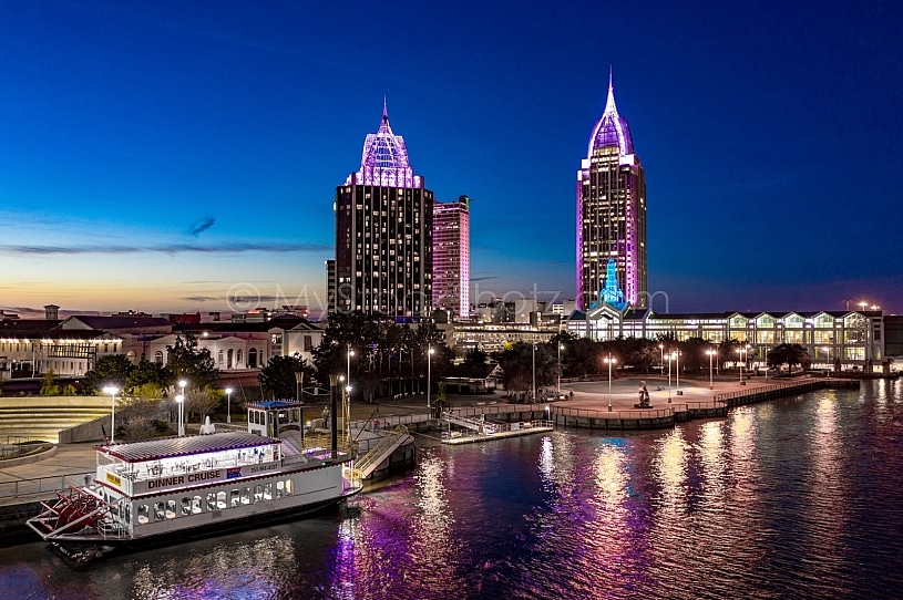 Vibrant night view of RSA BankTrust towers glowing purple over Mobile River, dinner cruise docked—couple will next go to the romantic wedding venue in Pensacola.