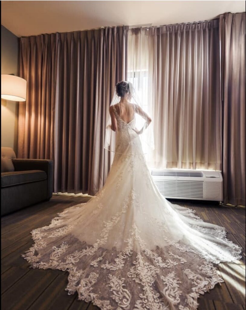 Bride in elegant lace wedding gown with cathedral train standing by sunlit window in hotel room, perfect for wedding venues in Pensacola FL.