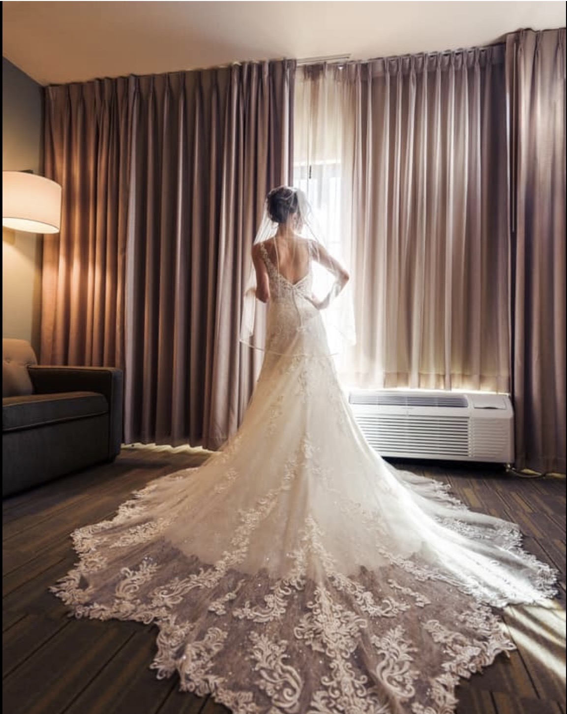 Bride in elegant lace wedding gown with cathedral train standing by sunlit window in hotel room, perfect for wedding venues in Pensacola FL.